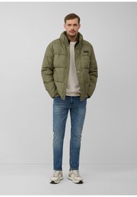 Olijfgroene pufferjack met geborduurd patroon, drukknoopsluiting en een logo-patch, gedragen over een lichte trui en gecombineerd met blauwe jeans en sneakers.