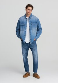 La giacca di denim presenta un design a gradiente blu, due tasche sul petto, chiusura con bottoni, abbinata a jeans di denim blu chiaro e scarpe marroni.