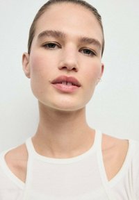 Primo piano di un modello che indossa un indumento bianco aderente con design a spalla scoperta. Pelle liscia con trucco sottile, che mette in evidenza labbra naturali e orecchini.