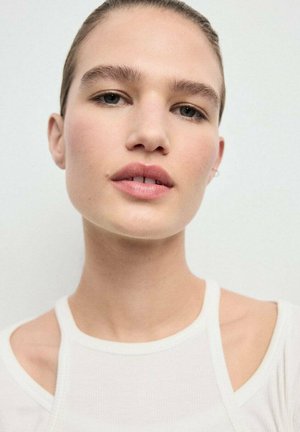 Primer plano de un modelo vistiendo una prenda blanca ajustada con diseño de hombros descubiertos. Piel suave con un maquillaje sutil, mostrando labios naturales y pendientes.