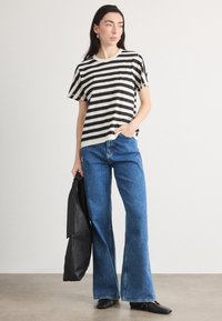 Cămașă cu dungi alb-negru, cu mânecă scurtă, asortată cu blugi din denim albastru cu picioare largi. Modelul ține o poșetă neagră. Texturi netede și croială lejeră.