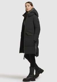 Schwarzer Puffermantel mit Kapuze, Reißverschluss vorne und seitlichen Taschen. Strukturierter Stoff, knielang, kombiniert mit schwarzen Schnürstiefeln.