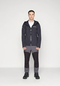 Zwarte waterdichte jas met een ritssluiting aan de voorkant en een capuchon, gecombineerd met grijze en zwarte broek. Gestructureerde stof met zichtbare logo-accenten.