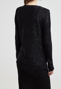 Svart stickad tröja med en texturerad design och metalliska accenter. Den har en rund halsringning och en lång siluett.