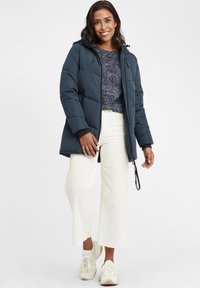 Marineblå polstret jakke med hætte, lynlåslukning og elastiske manchetter, parret med en spottet sweater og hvide brede bukser samt sneakers.