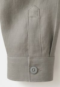 Poignet de manchette en tissu gris clair avec un seul bouton gris et un détail de bride en tissu cousu au-dessus de la couture du poignet.