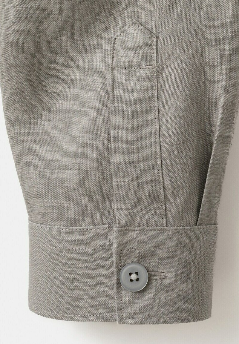 Poignet de manchette en tissu gris clair avec un seul bouton gris et un détail de bride en tissu cousu au-dessus de la couture du poignet.