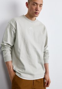 Cămașă Nike de culoare gri deschis, cu mânecă lungă, guler rotund și logo brodat pe piept, asortată cu pantaloni maro. Material din bumbac moale.