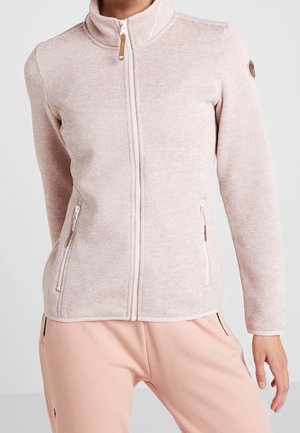 Jasnoróżowa polarowa kurtka na zamek błyskawiczny z wysokim kołnierzem i zapinanymi na zamek kieszeniami bocznymi, noszona przez osobę w brzoskwiniowych spodniach na jednolitym tle.