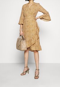 Vestido floral envolvente en mostaza con mangas de campana y dobladillo con volantes, combinado con un bolso beige y sandalias de tiras verde oliva.