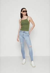 Aliejinių žalių prigludusių marškinėlių ir šviesiai mėlynų suplėšytų skinny džinsų derinys. Modelis vilki juodus saulės akinius ir batus baltais padais. Minimalistinis fonas.