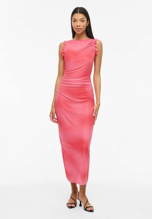 Femme debout, portant une robe midi rose sans manches avec des épaules froncées et des sandales à talons noires sur un fond blanc.