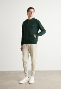 Felpa verde con cappuccio, tasca frontale e coulisse, abbinata a pantaloni della tuta beige chiaro e sneakers bianche. Stile semplice e taglio rilassato.
