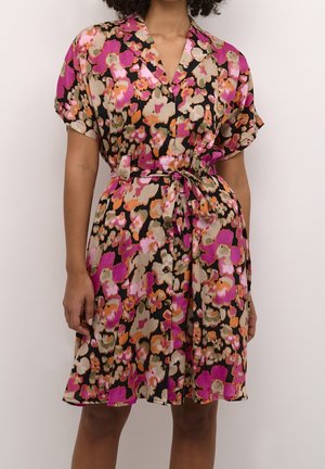 Vrouw draagt een ceintuur, korte mouwen jurk met een meerkleurig bloemenpatroon in roze, oranje, beige en zwarte tinten.