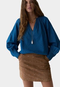 Blusa in cotone blu con scollatura a V profonda e maniche voluminose, abbinata a una gonna in suede marrone con decorazioni in argento.
