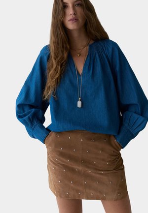 Blouse en coton bleu avec un décolleté en V profond et des manches volumineuses, associée à une jupe en daim marron ornée de paillettes argentées.