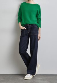 Grön stickad tröja, mörkblå vida jeans med kontrasterande sömmar, och vita sneakers med beige accenter.