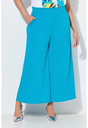 Weite türkisfarbene Hose mit Seitentaschen, getragen mit einem weißen gemusterten Oberteil und silbernen spitzen High Heels.