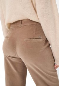 Bruine corduroybroek met een gladde textuur, voorzien van twee achterzakken en een klein metalen merkembleem, gedeeltelijk bedekt door een beige sweater.