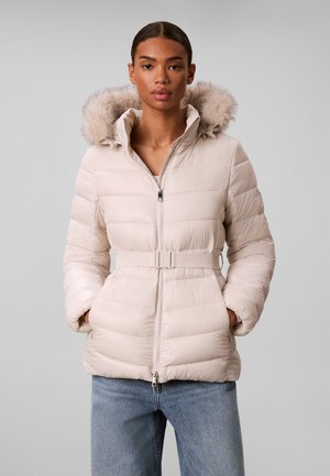 Femme portant une doudoune matelassée beige clair avec capuche bordée de fourrure et ceinture, debout les mains dans les poches, fond gris.