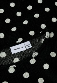Sort stof med tekstureret overflade, der har hvide prikker. Rund halsudskæring med synlige syninger. Mærket "name it" vises.