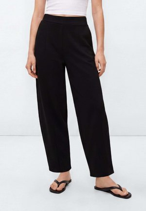 Personne portant un pantalon large noir taille haute et des tongs noires, associé à un top blanc court, debout devant un fond uni.