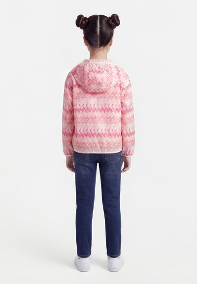 Enfant vu de dos portant un jean bleu, des baskets blanches et une veste à capuche rose à motifs, avec les cheveux coiffés en deux chignons.