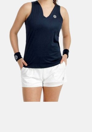 Woman wearing a sleeveless navy blue sports top, white shorts, and matching navy wristbands with logos, hands in pockets.