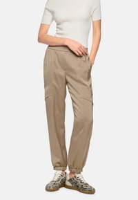 Beige tapered cargo pants with elastic cuffs, featuring two front pockets and a smooth texture, paired with patterned sneakers.