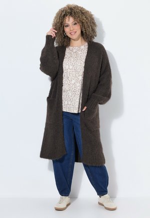 Kvinde med krøllet hår iført en lang, mørkebrun cardigan, mønstret beige skjorte, blå jeans og hvide sneakers, står og smiler.