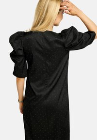 Rochie neagră cu mâneci bufante, decorată cu mici puncte metalice. Textură netedă, cu o croială ajustată, prevăzută cu închidere la spate.