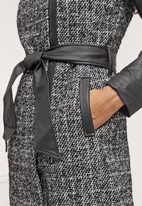Schwarz-weißer gemusterter Jumpsuit mit taillierten Design, Lederelementen, einem gebundenen Gürtel und Seitentaschen, ausgestattet mit einer strukturierten Stoffoberfläche.