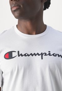 Camiseta de algodón blanca con un gran logo negro de "Champion" con un acento rojo, cuello redondo y mangas cortas. Textura suave, diseño casual.