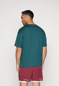 Sötétzöld, rövid ujjú póló, amely puha anyagból készült, és kerek nyakkivágással rendelkezik. Bordó sportnadrággal párosítva, amelyen fehér felirat látható.