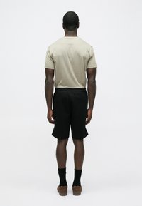 Bézs színű, pamut póló, fekete rövidnadrág, texturált, rugalmas derékpánttal, fekete zoknival és barna cipővel viselve, hátulról nézve.