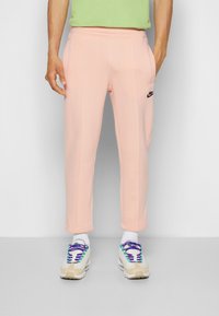 Světle růžové tepláky s černým logem, hladká textura, elastický pas a dvě boční kapsy, doplněné multibarevnými sportovními botami.