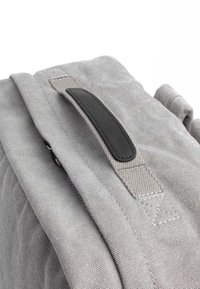 Grau Stoff-Rucksack mit strukturierten Oberflächen, ausgestattet mit einem schwarzen Leder-Tragegriff und Reißverschlussbeschlägen. Sichtbare genähte Akzente.