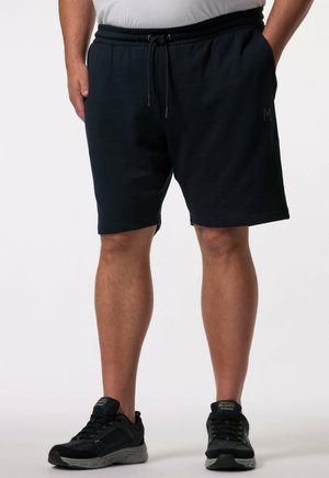 Mann trägt schwarze Sportshorts mit Kordelzug und schwarze Sneakers, steht mit den Händen in den Taschen vor weißem Hintergrund.