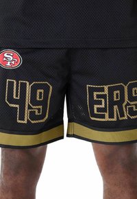 Pantalones cortos deportivos de malla negra con detalles dorados, un dobladillo inferior ancho y grandes números "49" y "ERS" en la parte delantera, con un logo de SF en el lateral.