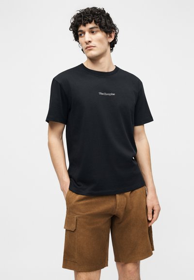 Homme aux cheveux bouclés portant un t-shirt noir à manches courtes avec un petit texte blanc « The Kooples » et un short cargo marron, les mains dans les poches, regardant vers la gauche.