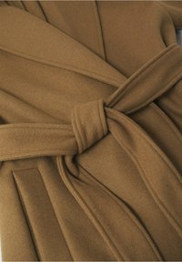 Gros plan sur un manteau en laine marron avec une ceinture en tissu nouée et des coutures visibles sur un tissu à texture douce.
