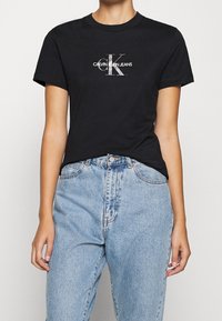 Camiseta de algodón negra con mangas cortas y cuello redondo, con el logotipo blanco de Calvin Klein Jeans en el frente. Combinada con jeans azul claro.