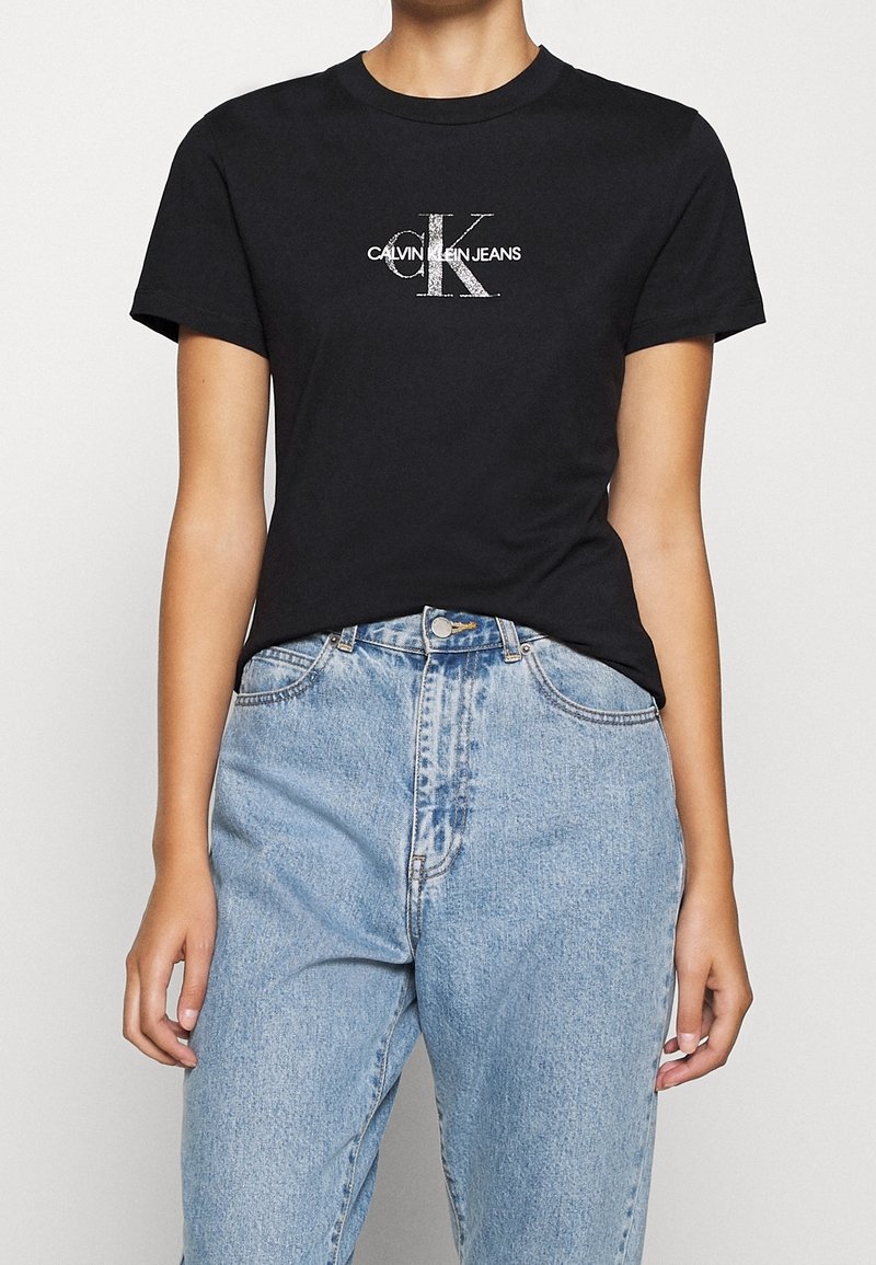 Camiseta de algodón negra con mangas cortas y cuello redondo, con el logotipo blanco de Calvin Klein Jeans en el frente. Combinada con jeans azul claro.