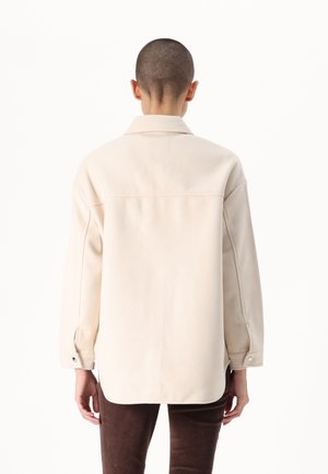 Person with short hair wearing a loose beige buttoned shirt jacket and dark brown corduroy pants, viewed from behind.