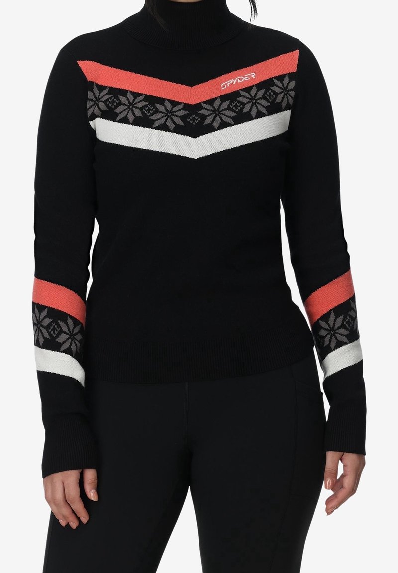 Pull à col roulé noir avec motif flocon de neige rouge, blanc et noir sur la poitrine et les manches, porté par une personne avec les mains détendues le long du corps.