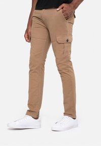 Kaki cargobroek gemaakt van stevig stof, met zijzakken, een slanke pasvorm en contrasterende stiksels. Draagt witte sneakers.