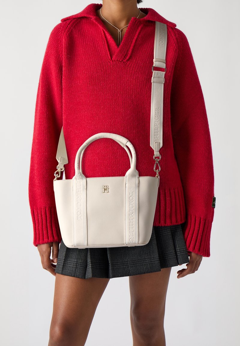 Femme portant un pull en maille rouge et une jupe plissée noire porte un sac à main en cuir blanc avec une bandoulière ajustable.