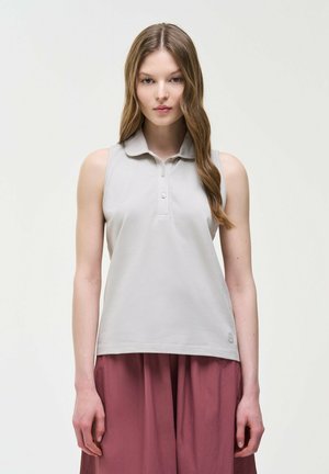 Mladá žena s dlhými vlnitými vlasmi, oblečená v bezrukávovom svetlosivom top s golierom a voľných bordových nohaviciach na jednofarebnom bielom pozadí.