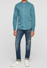 Chemise turquoise à boutons avec col mao, associée à un jean en denim bleu déchiré et des baskets blanches.