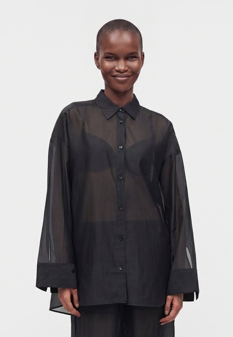 Personne souriante portant une chemise noire ample et transparente à boutons par-dessus un soutien-gorge noir, se tenant devant un fond uni clair.